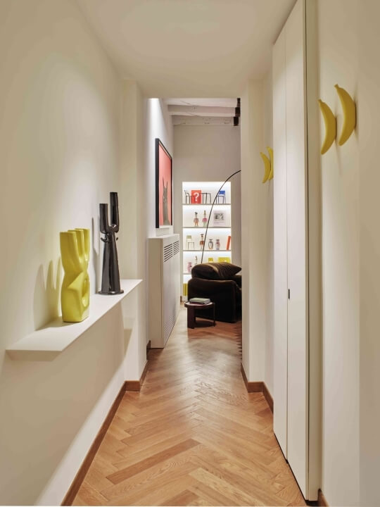 A modern hallway with white walls, wooden herringbone floors, yellow vases, a black sculpture, and a doorway to another room.