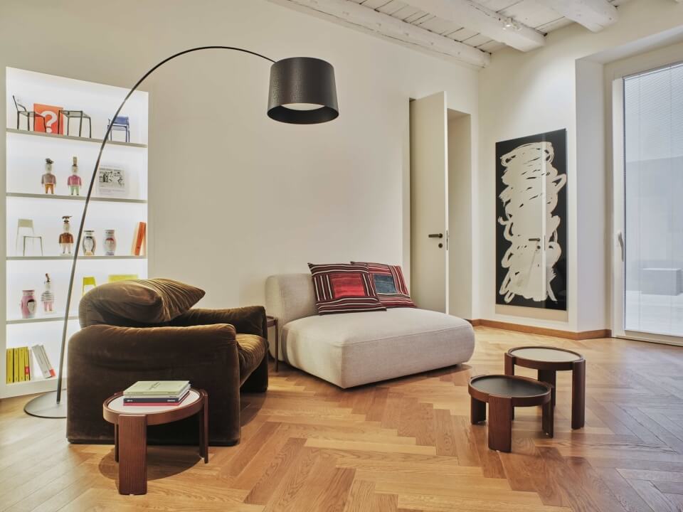 Soggiorno moderno con pareti bianche, pavimento in legno, poltrona marrone, chaise longue beige e lampada curva.