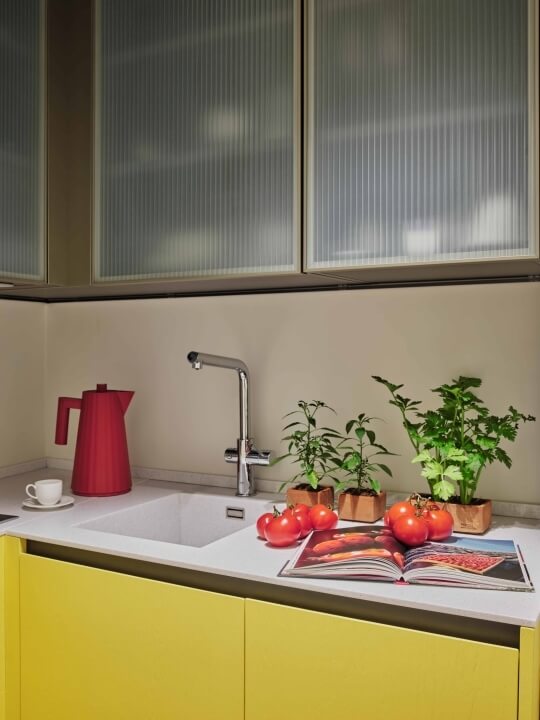 Bright kitchen with white countertop, yellow cabinets, red coffeepot, potted plants, tomatoes, and an open cookbook.