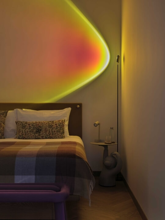 A cozy bedroom with a bed, nightstand, lamp, and purple chair, featuring a warm glow and rainbow shadows on white walls.