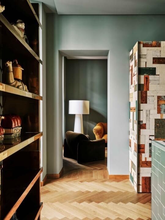 A cozy hallway with light blue walls, wooden zigzag floor, shelves of decor, and a doorway to a room with an armchair and ottoman.