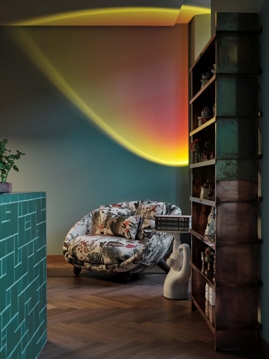 A modern room features a vibrant yellow-orange wall installation, a round chair, a bookshelf, and minimal decor.