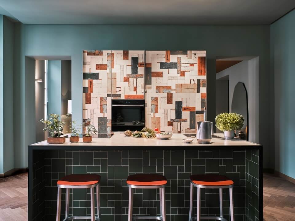 Modern kitchen with a unique tile backsplash, central island, decorative plants, and arched doorway in light blue-gray walls.