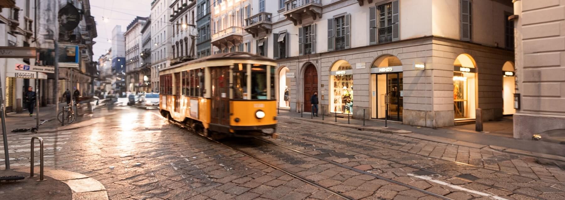 A yellow tram moves down a wet brick street, surrounded by urban buildings, under a grayish-white sky at dusk.