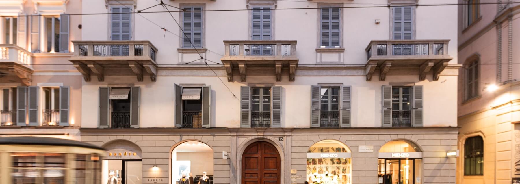 Urban building with beige facade, gray shutters, arched doors, small balconies, and a blurred trolley car in motion.