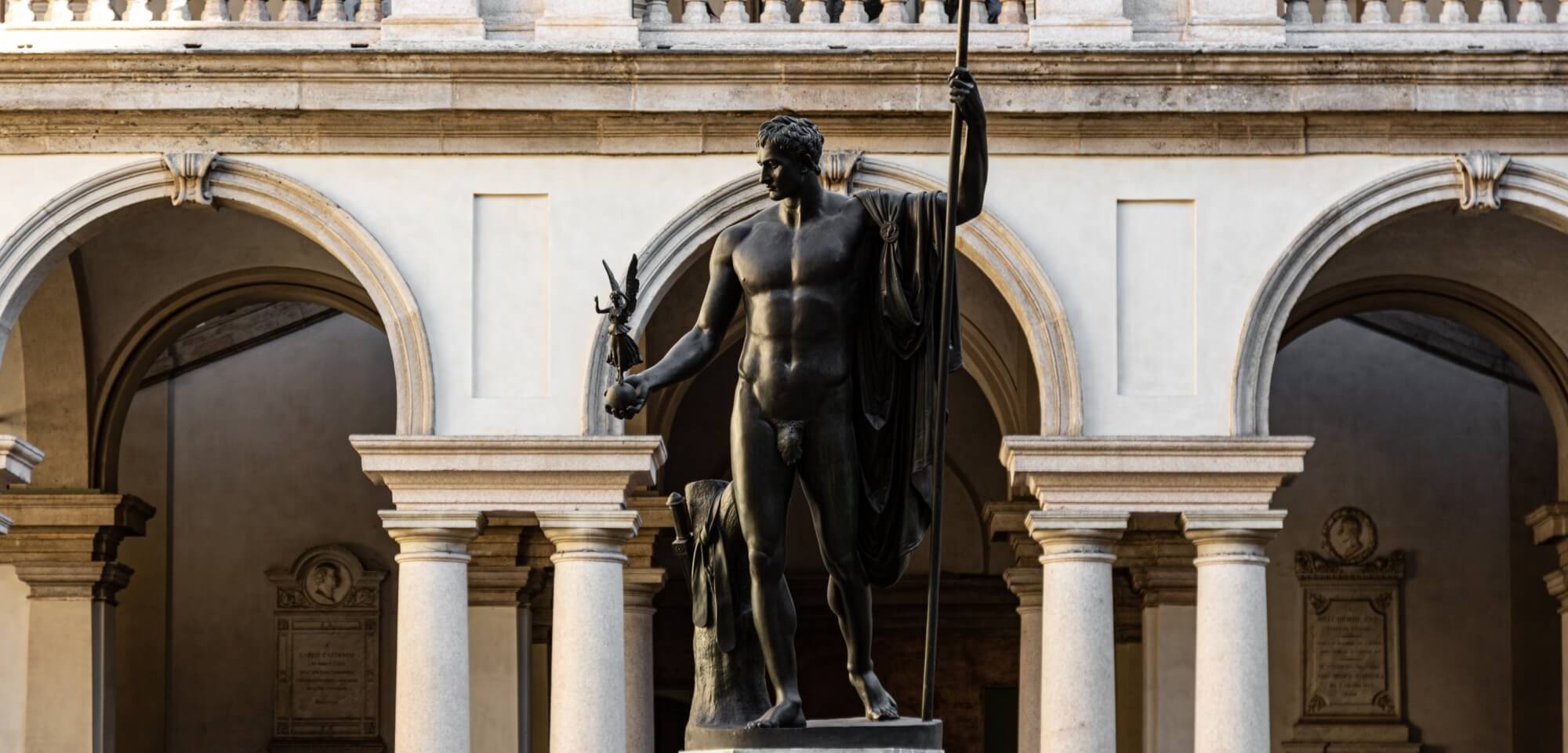 Statua di un uomo muscoloso con lancia e uccello, davanti a un arco ornato in pietra bianca. Atmosfera di grandezza storica.