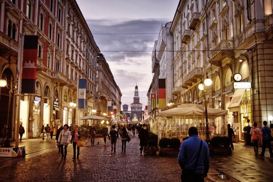 Bustling city street with ornate buildings, cobblestones, people with bags and umbrellas, and a distant clock tower.