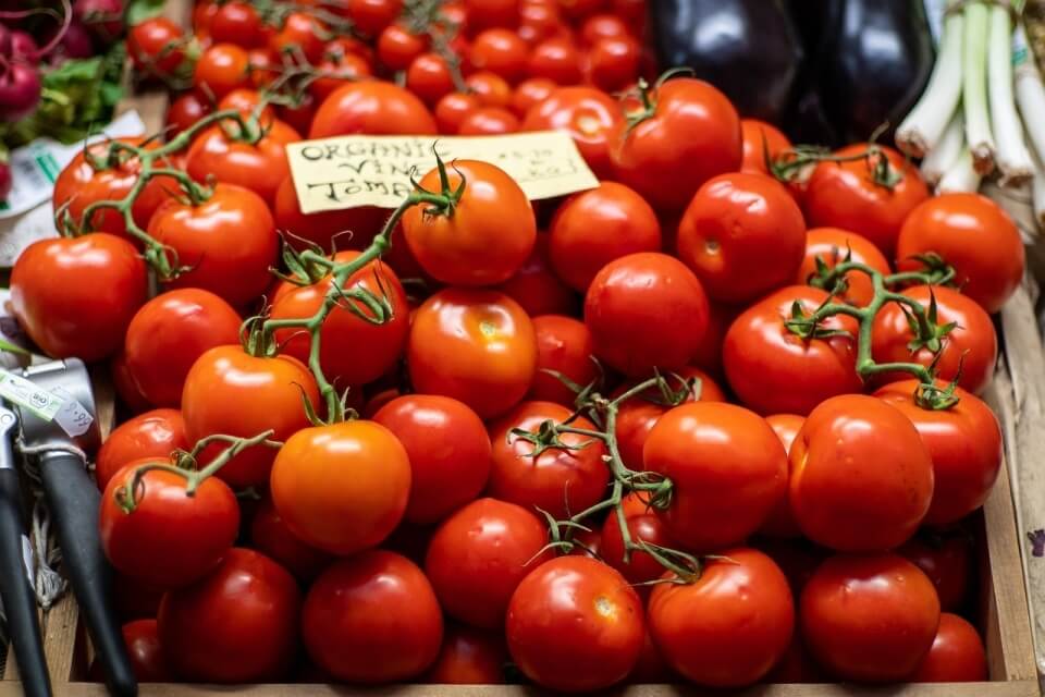 Wooden crate filled with red organic vine tomatoes, labeled with a handwritten note, surrounded by other fresh vegetables.