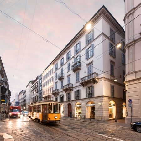 Historic yellow and brown streetcar on a cobblestone street in Milan, with buildings and a light pink-blue sky.