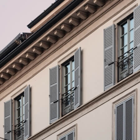 Multi-story building with cream facade, dark roof, blue shutters, and ornate moldings under an overcast sky.