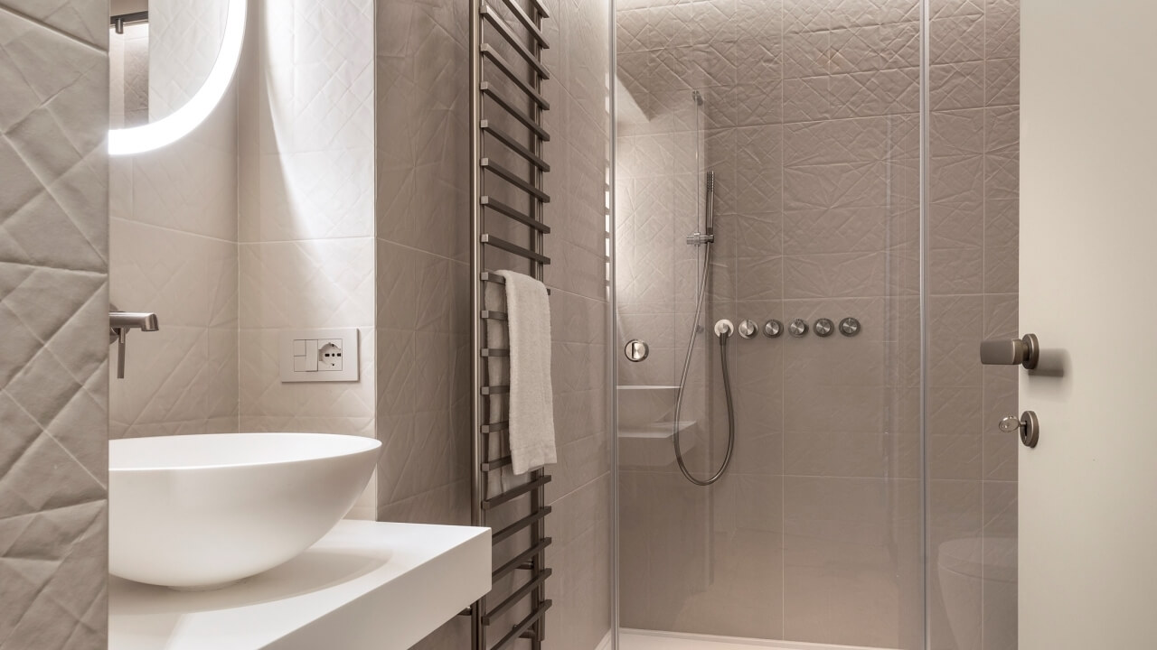 Modern bathroom with a white bowl sink, circular mirror, towel rack, and glass shower door, featuring beige tiled walls.