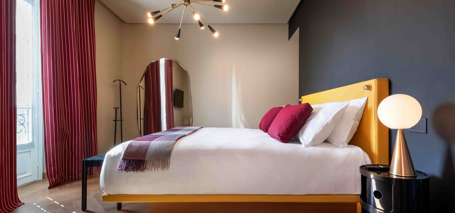 Modern bedroom featuring a double bed with a yellow headboard, red and purple blanket, black nightstand, and gold chandelier.