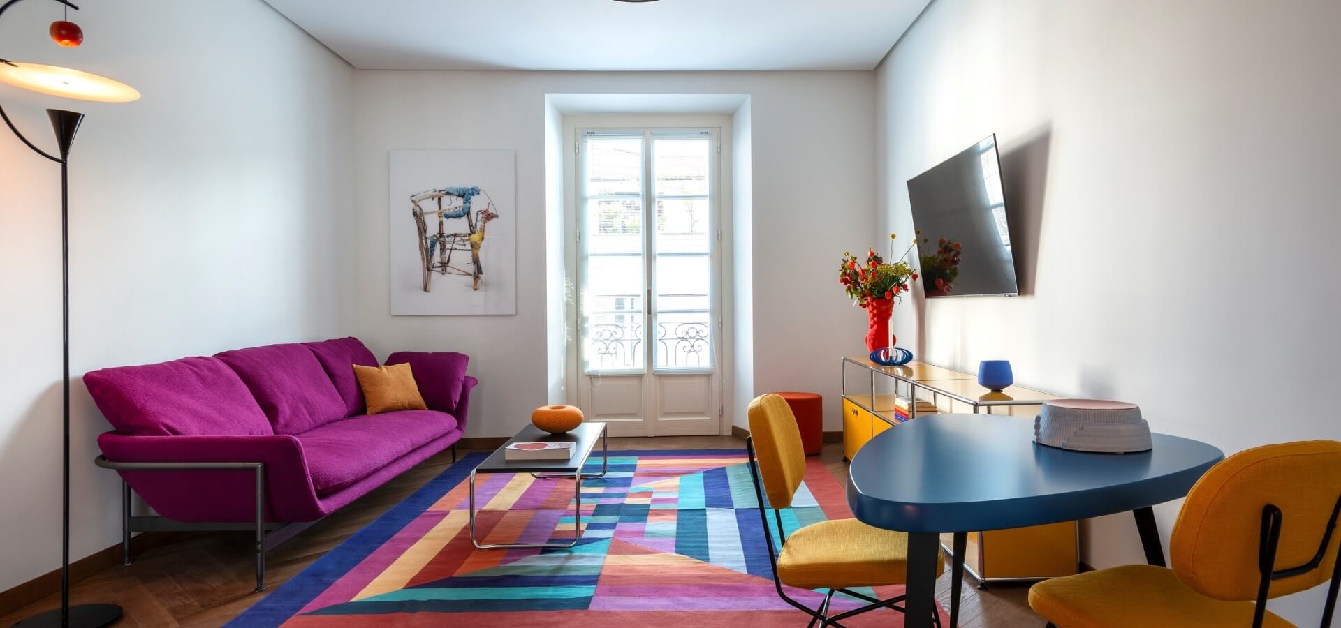 Modern living room with purple couch, blue table, yellow chairs, and vibrant decor, illuminated by natural light from a large window.