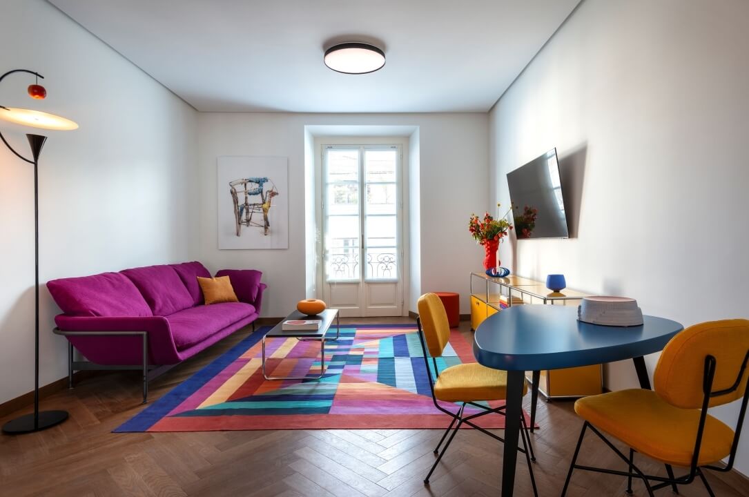 Modern living room with a purple couch, blue table, yellow chairs, multicolored rug, and bright natural light.