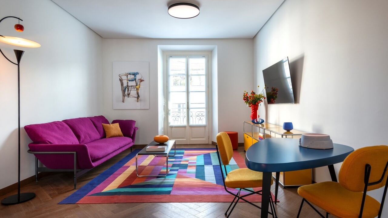 Modern living room with a purple couch, yellow chairs, blue table, colorful rug, and large window, exuding sophistication.