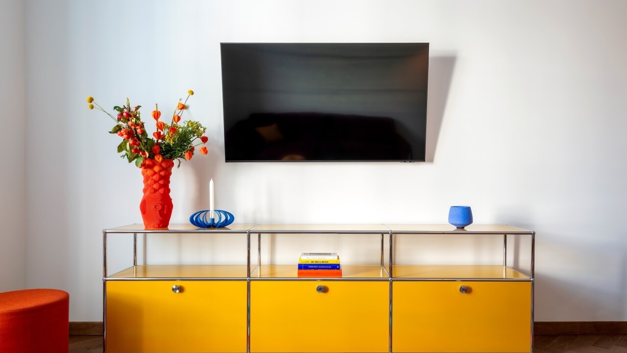 Modern living room with yellow and silver entertainment stand, flat-screen TV, vibrant flowers, and hardwood floors.