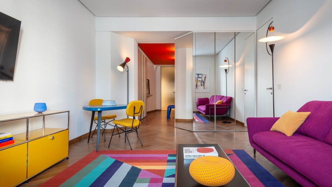 Modern living room with a purple couch, brown coffee table, multicolored rug, and vibrant decorative elements.