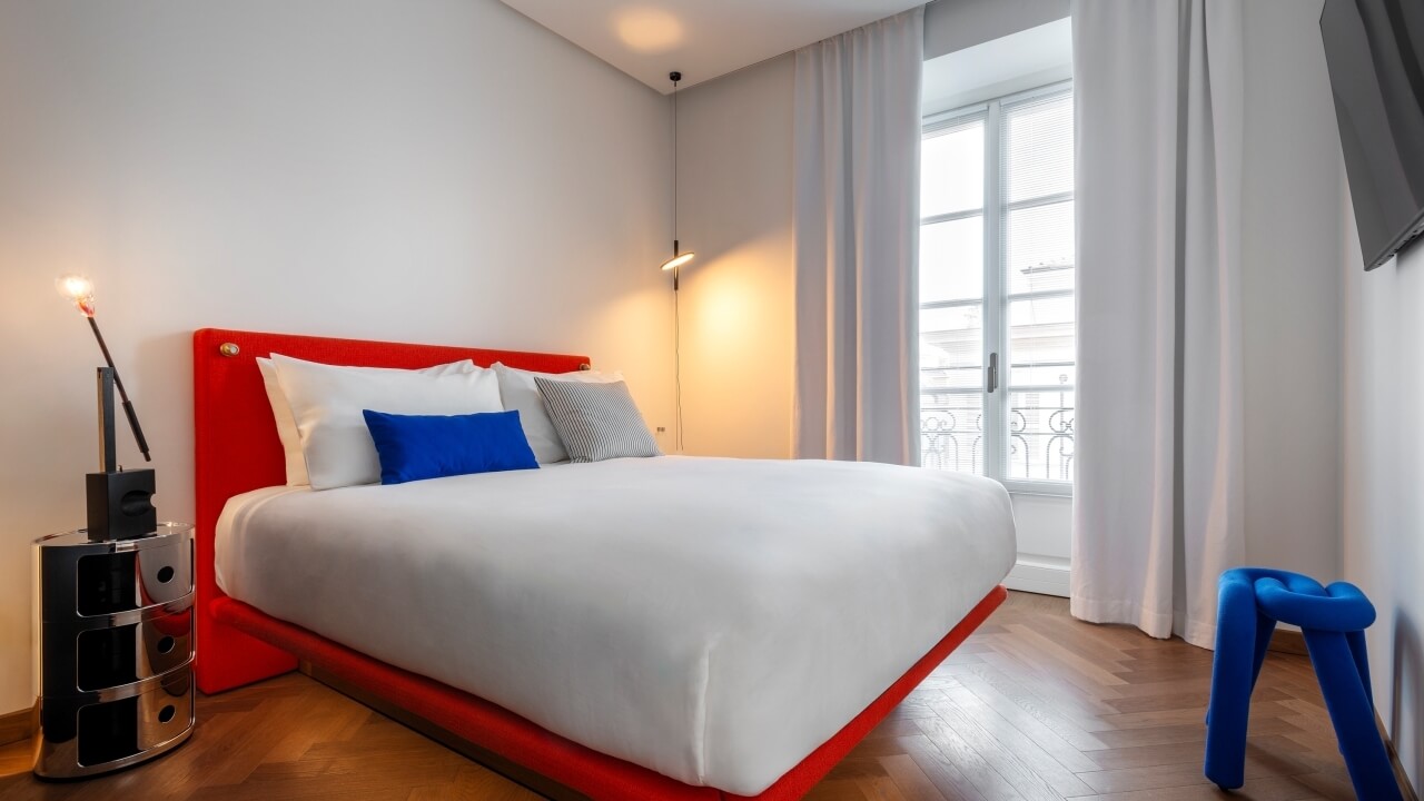 Modern bedroom with red bed, white duvet, sleek nightstand, gray curtains, blue stool, and mounted TV, exuding sophistication.