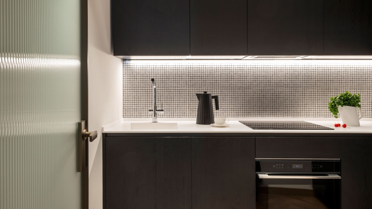 Modern kitchen with dark cabinets, white countertop, sink, greenery vase, and tomatoes against a black tile backsplash.