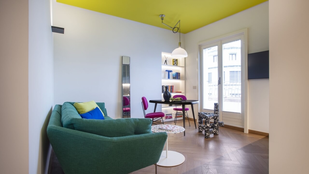 Modern living room with yellow ceiling, white walls, green couch, black dining table, and purple chairs, filled with natural light.