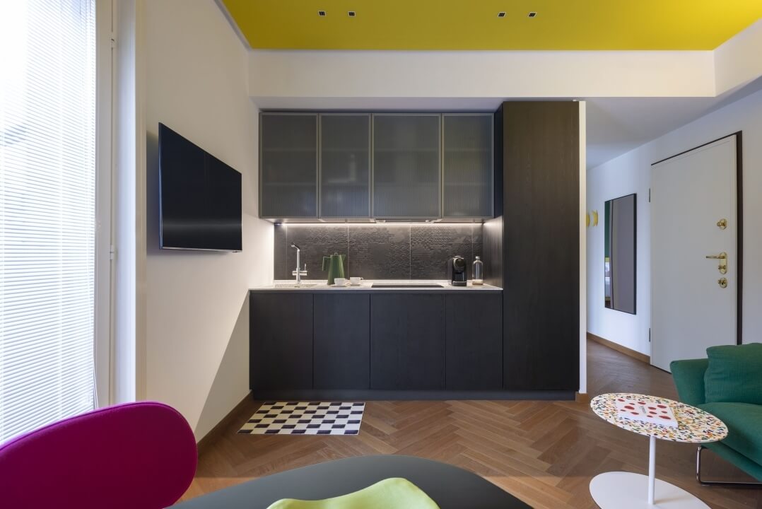 Spazio abitativo moderno con cucina, pareti bianche, soffitto giallo, sedia viola e divano verde, atmosfera accogliente.