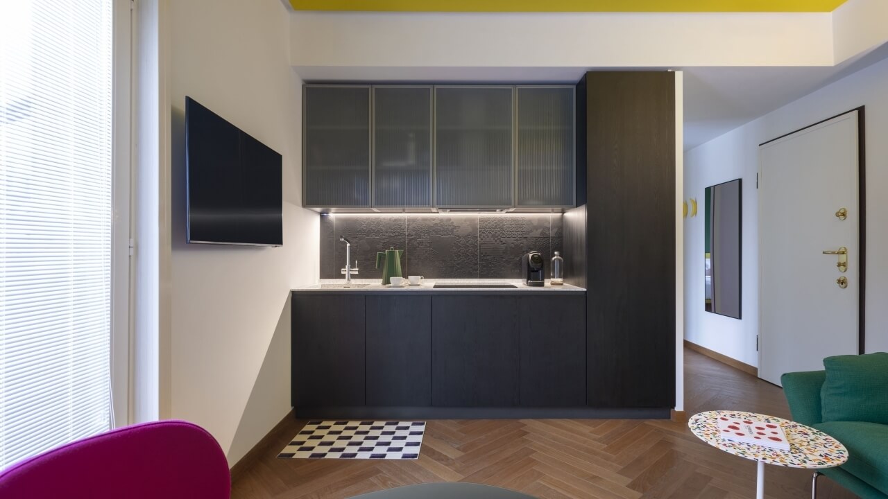 Modern kitchen and living room with neutral tones, dark cabinets, pink chair, and yellow accent wall, emphasizing simplicity and elegance.