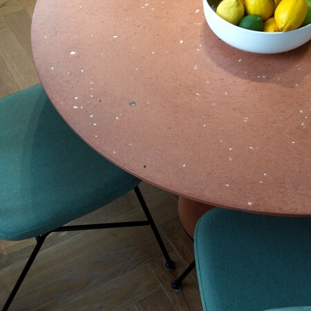 Top-down view of a terracotta table with white speckles, two teal chairs, and a bowl of lemons and limes on a wooden floor.