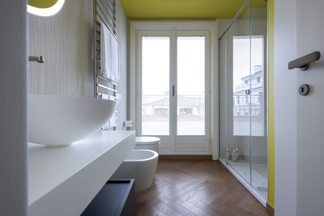 Bagno moderno con lavabo bianco, wc, doccia, pareti gialle e pavimento in legno chiaro, illuminato da una grande finestra.