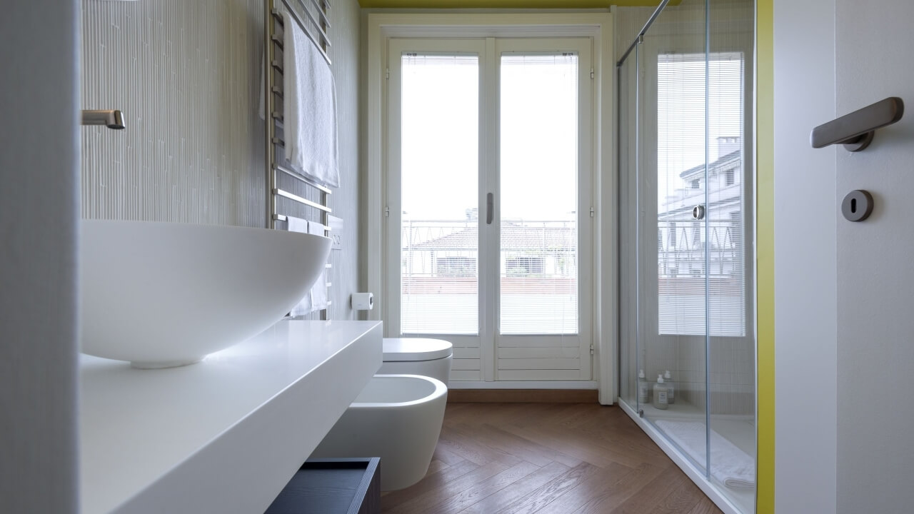 Bagno moderno minimalista con vanità bianca, doccia in vetro, pavimento in legno e vista esterna attraverso una porta.