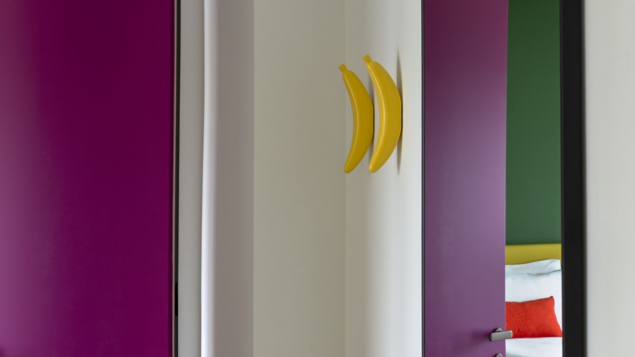 Interno vivace con porta verso una camera da letto, pareti bianche, pannello magenta e decorazioni di banane gialle.