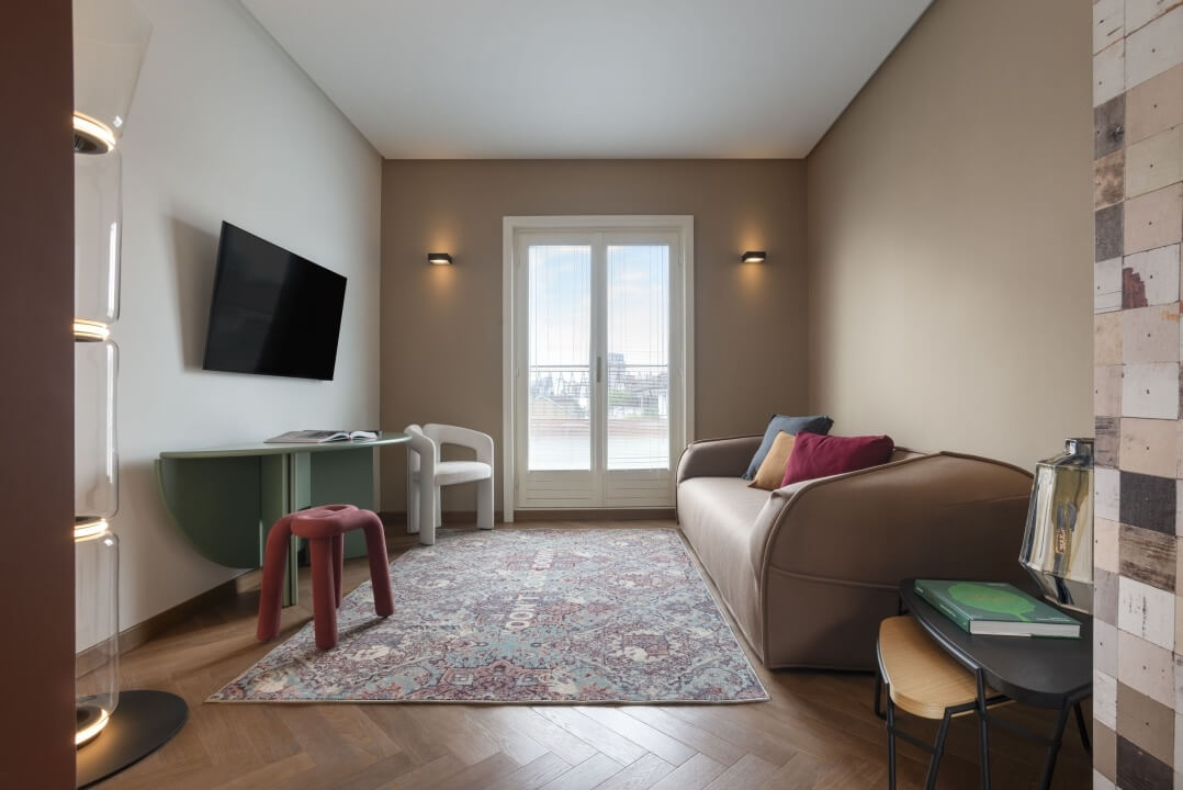 Salotto accogliente con divano beige, TV nera, tavolo in legno, sedia bianca, tappeto colorato e luce naturale dalla finestra.