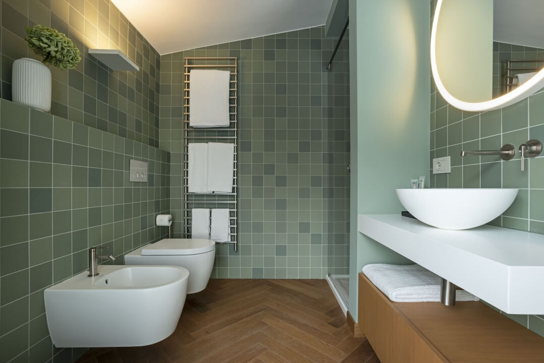 Bagno moderno con due wc a parete, lavabo, specchio retroilluminato e piastrelle verdi, design spazioso e funzionale.