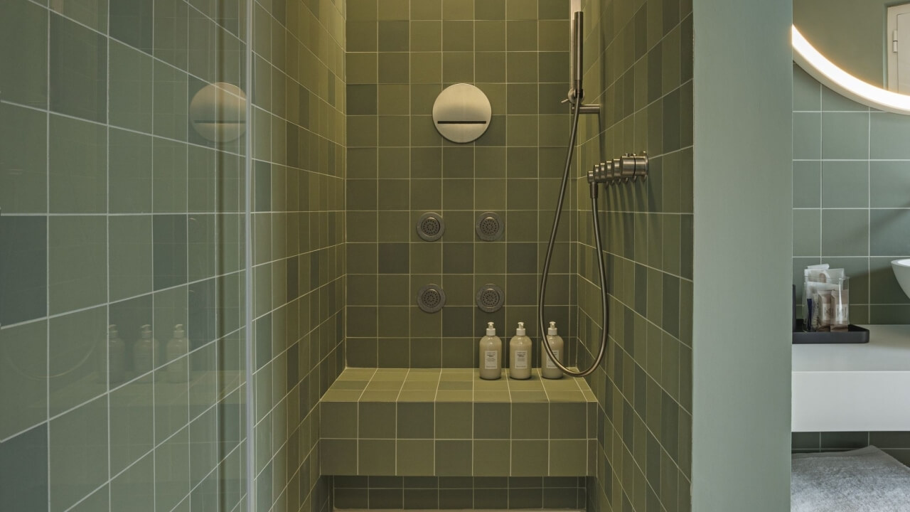 Modern bathroom featuring a green-tiled shower stall with three shower heads, a bench, and toiletries on a sink.