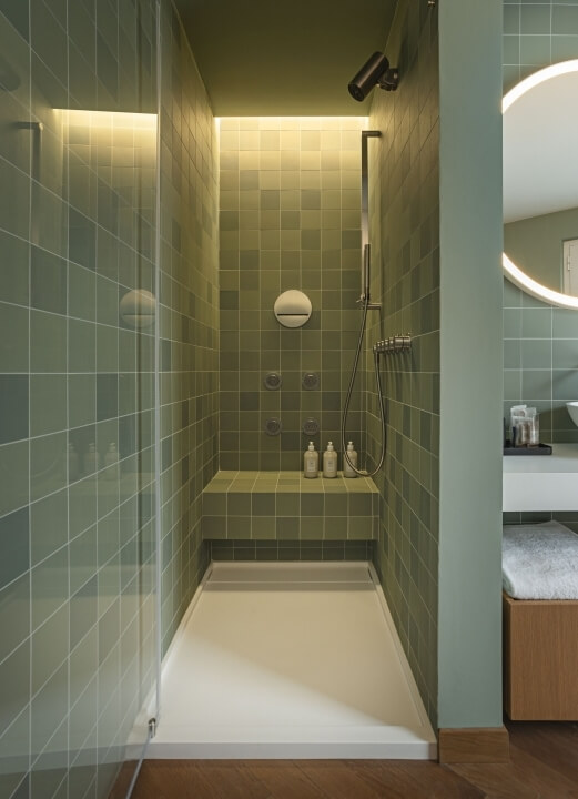 Bagno moderno con doccia a vetri, panchina bianca, specchi rotondi e pareti verde chiaro. Atmosfera pulita e funzionale.