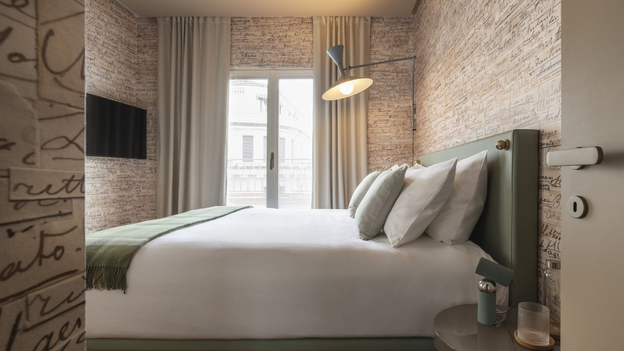 Camera d'hotel serena con letto bianco, testata salvia, lampada, e luce naturale che entra da una grande finestra.