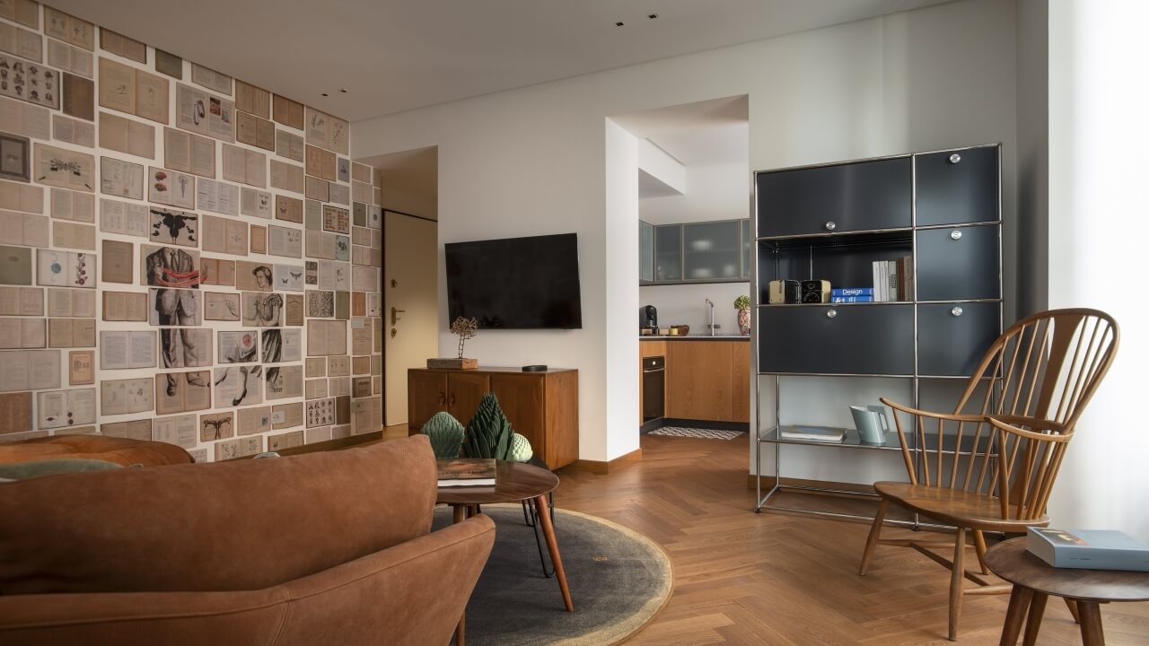 Modern living room with brown couch, collage wall, gray rug, wooden chair, and kitchen area in the background. Cozy atmosphere.