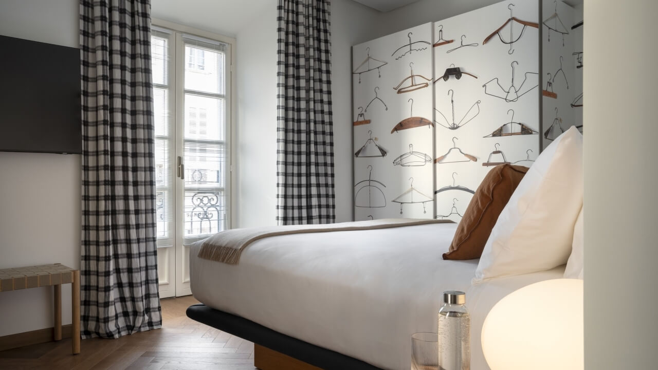 Modern bedroom with a large bed, unique coat hanger wall, window with plaid curtains, mounted TV, and warm lamp glow.