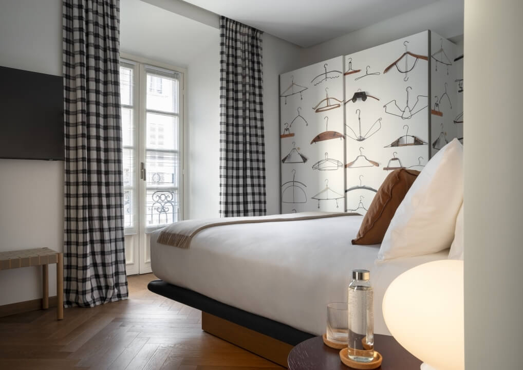 Camera poco illuminata con letto bianco, comodino, bicchieri d'acqua, schermo con appendiabiti e tende a quadri.