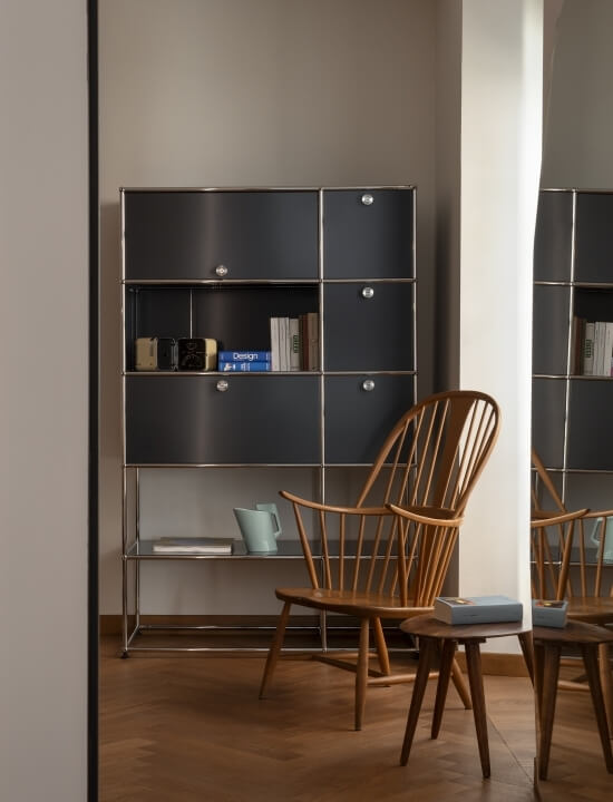 Una stanza con una libreria metallica, due sedie in legno, un tavolo rotondo e una finestra con tende bianche.