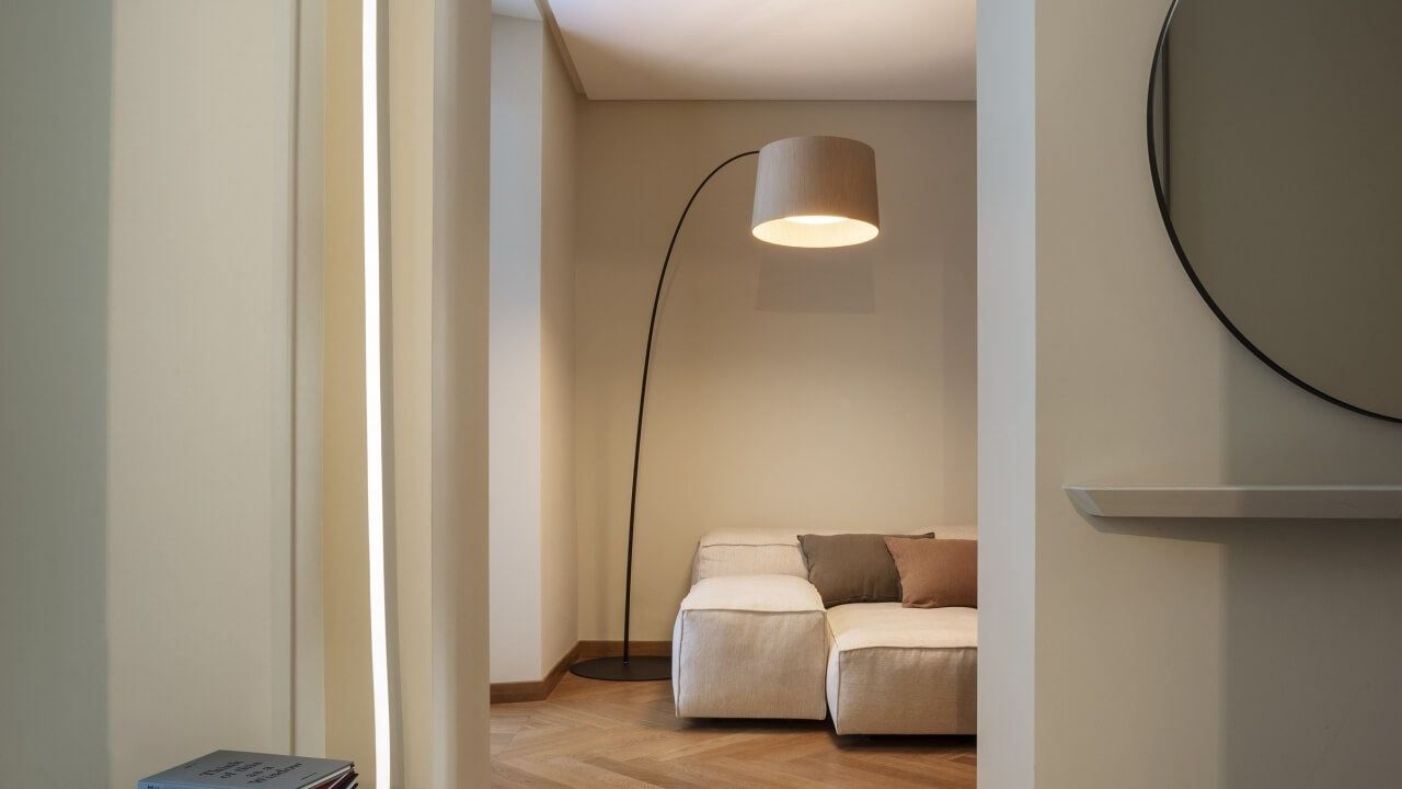 A cozy modern room with a black floor lamp, beige couch, round mirror, and small bookshelf against light beige walls.