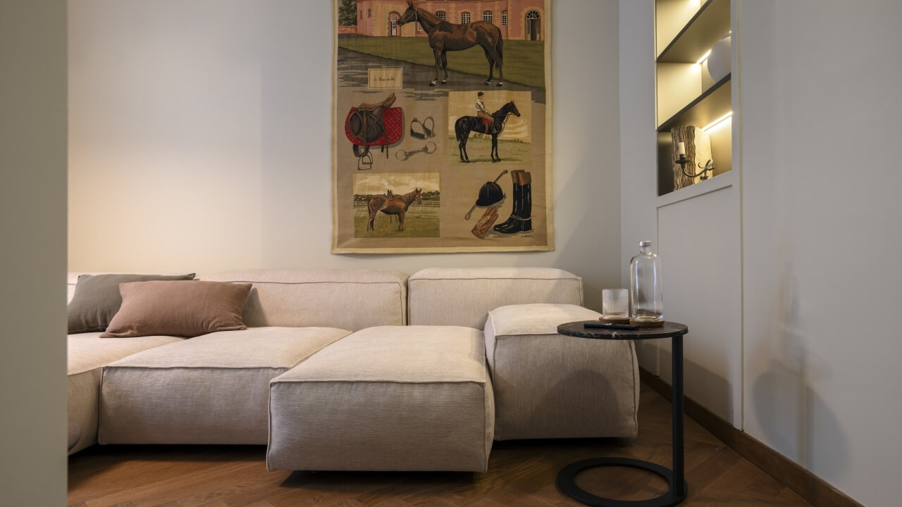 Cozy room corner with a beige sectional sofa, side table, tapestry of horse equipment, and decorative shelf.