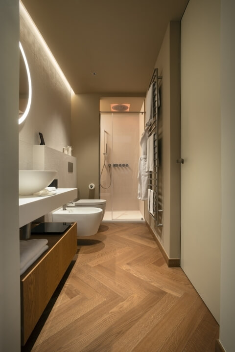 Bagno moderno ben illuminato con doccia, WC e lavabo, pareti beige e pavimento in legno a spina di pesce.