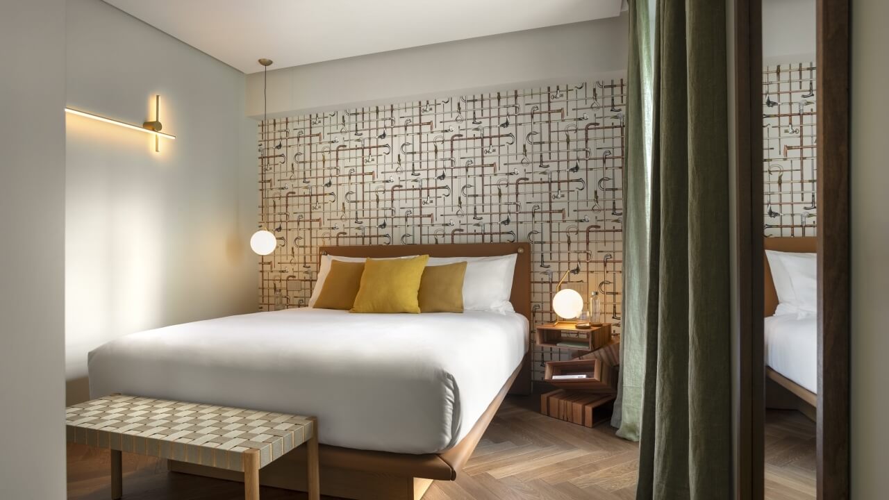 Modern hotel room featuring a bed with a white comforter, nightstand, patterned bench, and unique wall design.