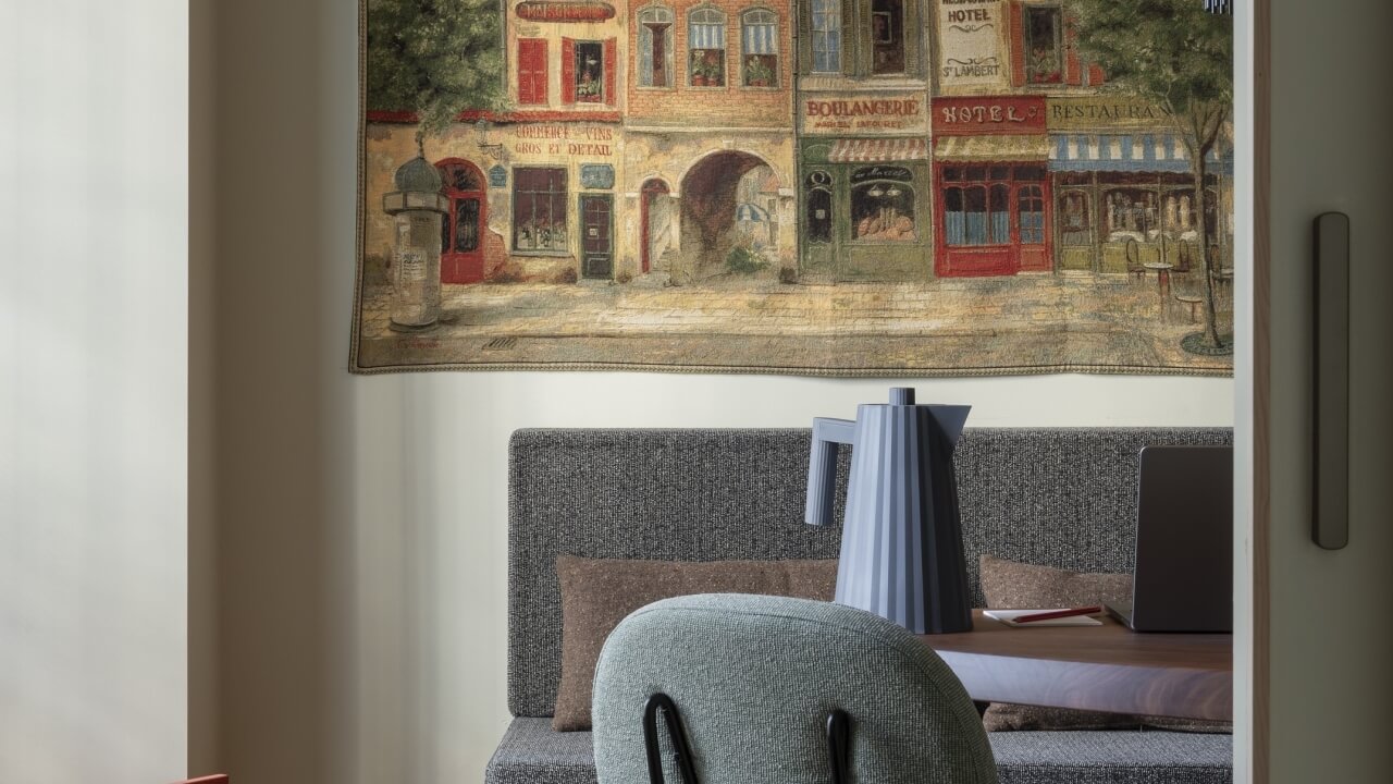 Interior seating area with a gray bench, green chair, round table, and a tapestry depicting a charming European street scene.