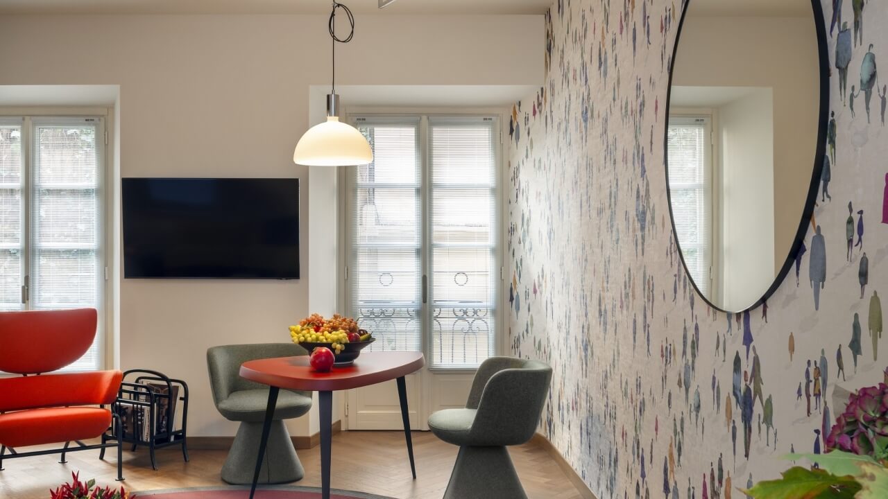 Modern living room with colorful decor, dining nook, orange chair, patterned wallpaper, and natural light from windows.