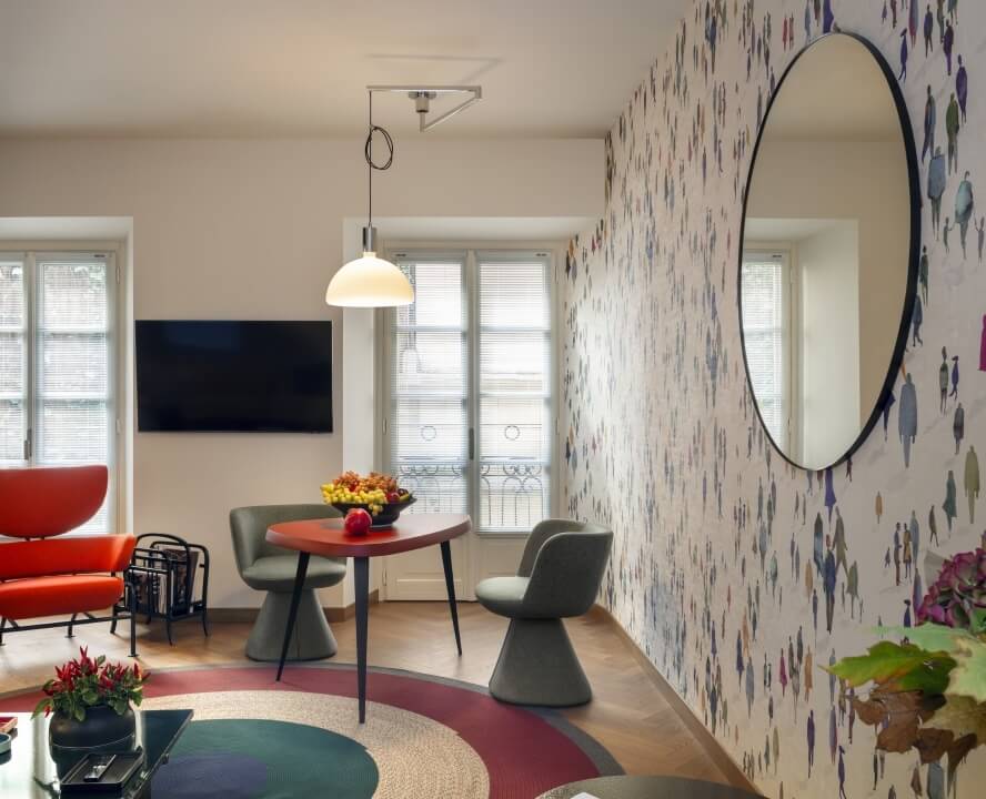 Soggiorno moderno con area pranzo, specchio rotondo, tappeto colorato, tavolo con frutta e piante, atmosfera accogliente.