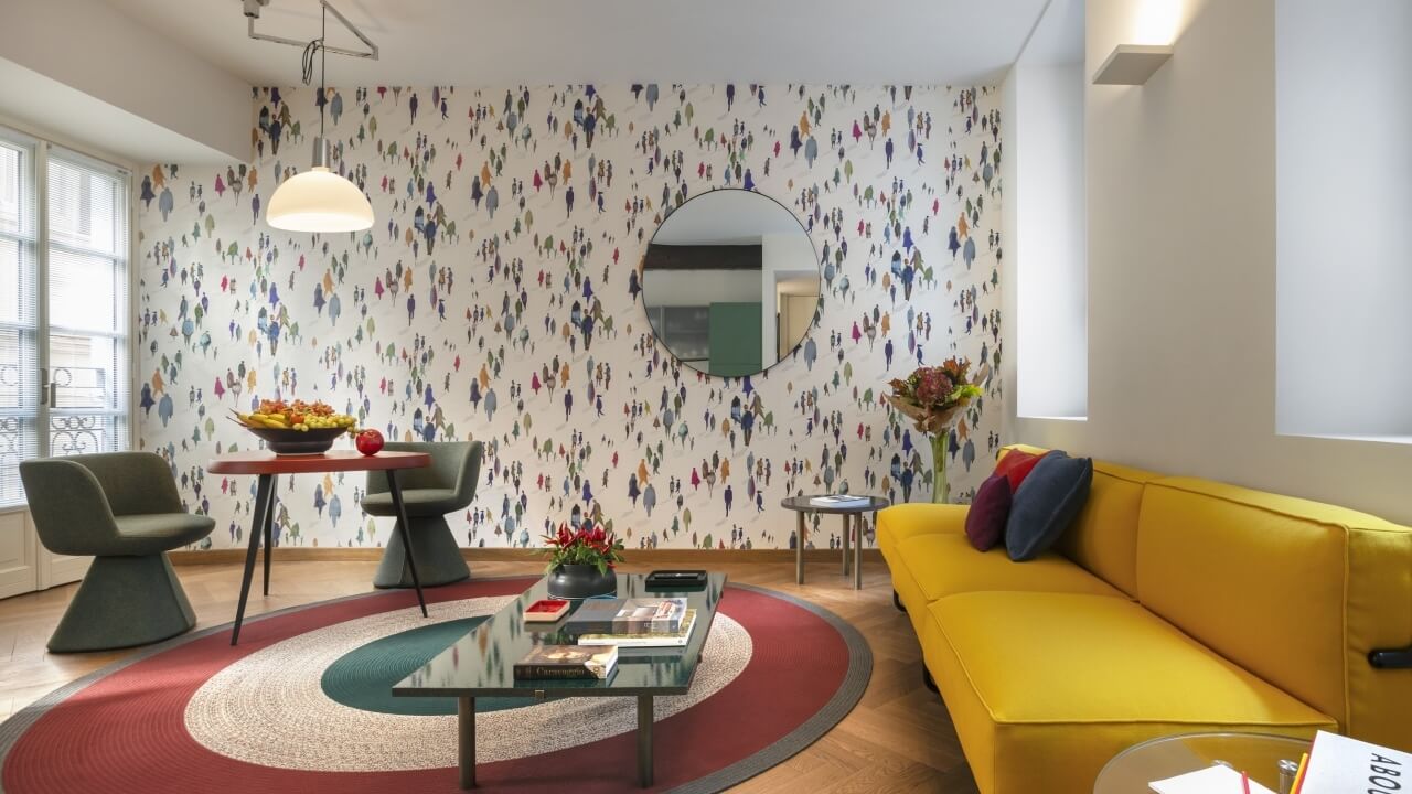 Modern living room with a yellow couch, colorful pillows, glass coffee table, and a cozy dining area with natural light.