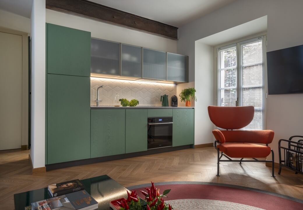 Cucina moderna con armadi verdi e soggiorno minimalista con sedia arancione, tavolino e luce naturale.