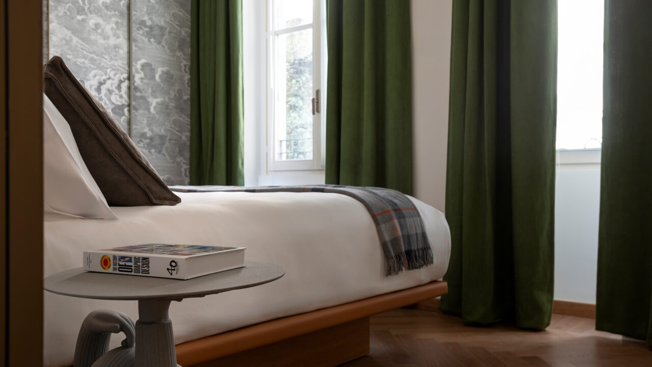 Serene bedroom with a white duvet bed, gray plaid blanket, round table with a book, and green curtains letting in natural light.