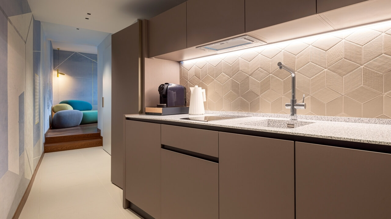 Modern kitchen with light brown cabinets, hexagonal backsplash, stainless steel sink, and a staircase with colorful cushions.